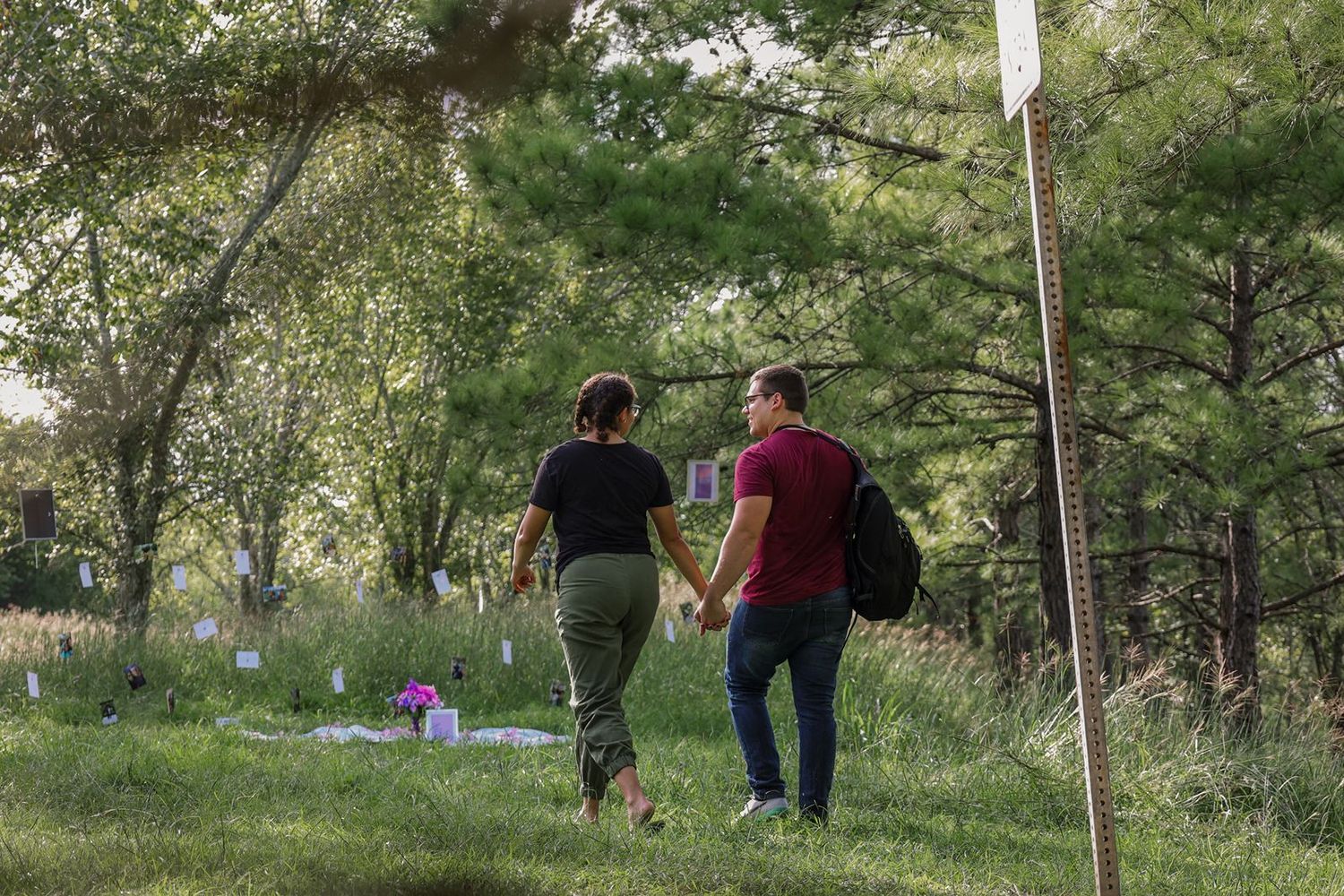 Couple holding hands walking through a green cemetery with flower memorials among trees.