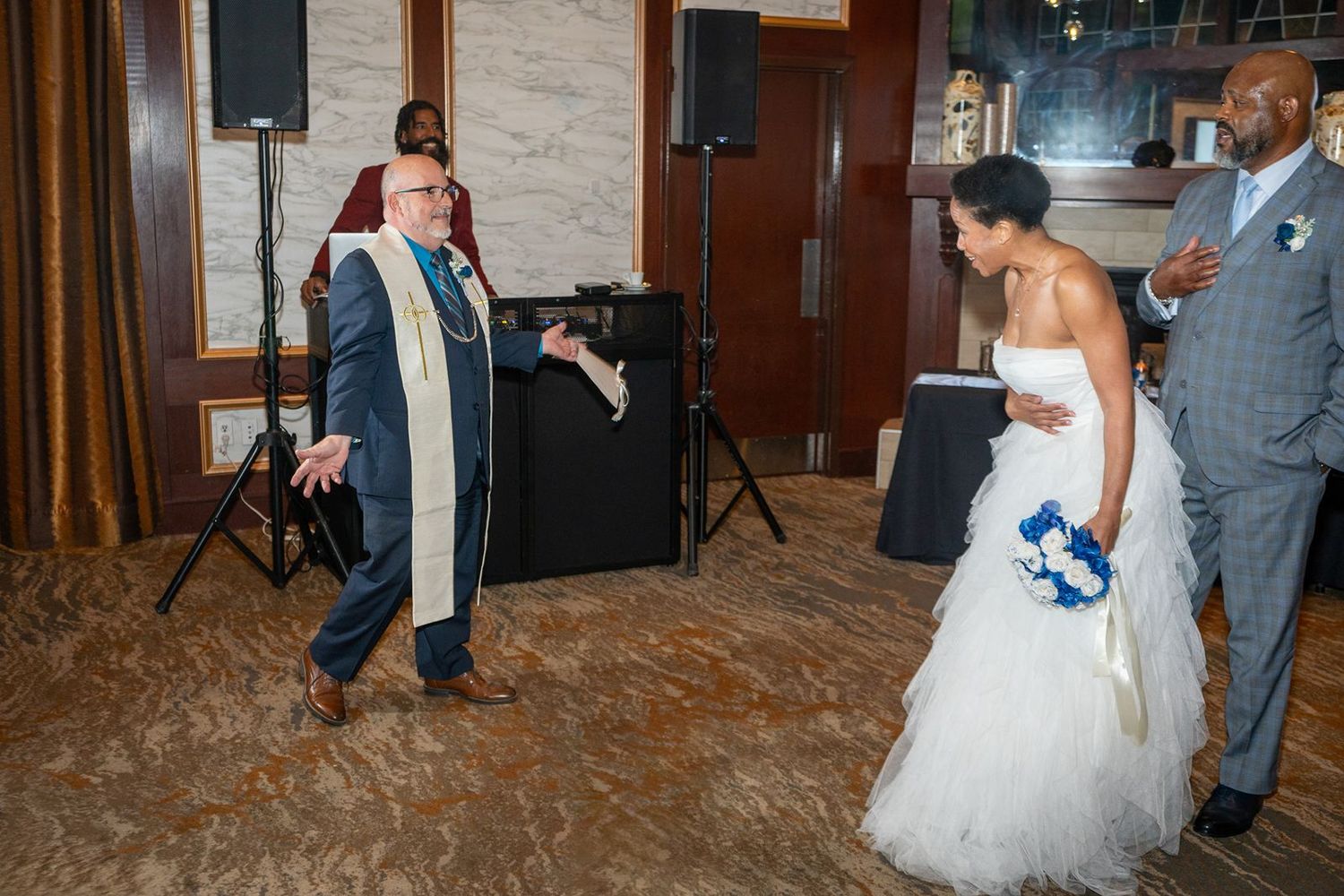 Wedding officiant dances energetically as bride in white gown and groom look on at reception.