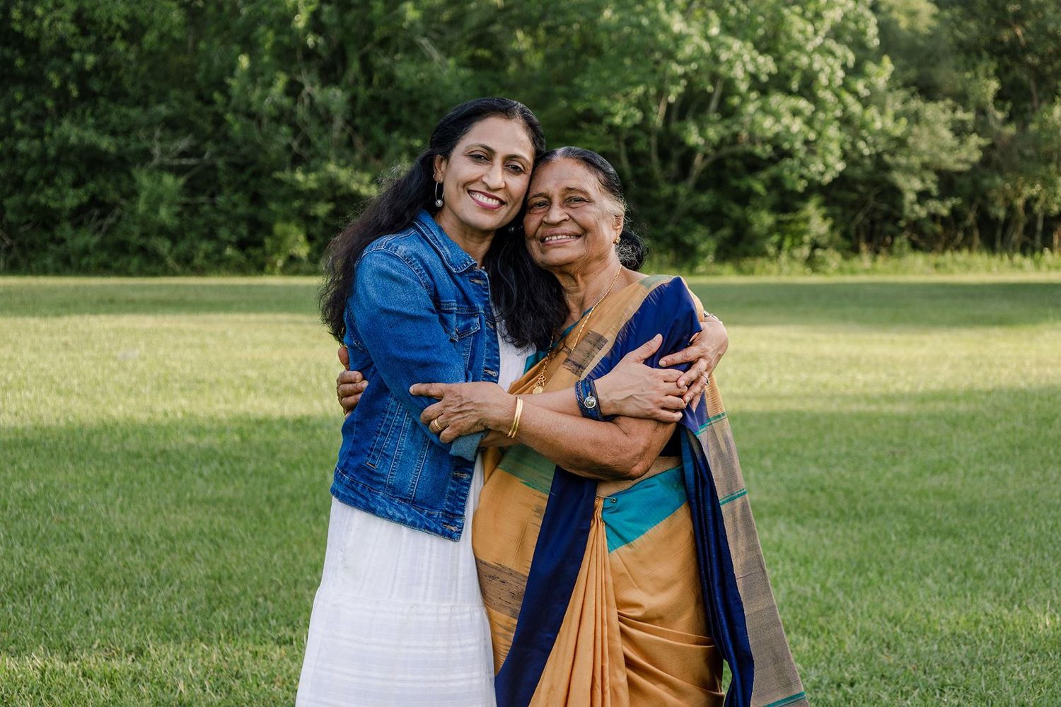 Two people embrace while smiling outdoors on a grassy lawn, one wearing a blue denim jacket and one in a gold sari.