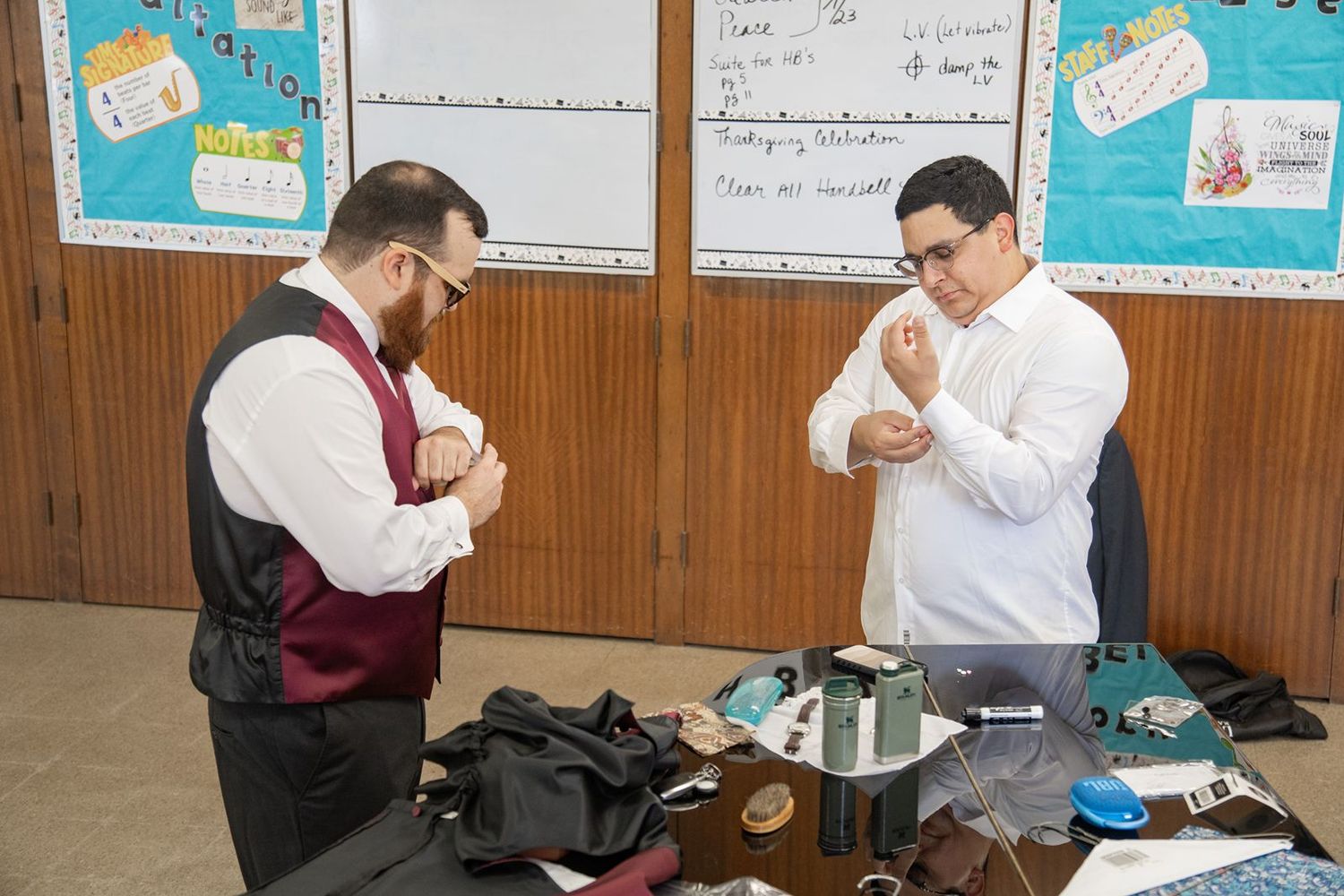 Two groomsmen prepare their wedding attire in a room decorated with colorful student artwork.