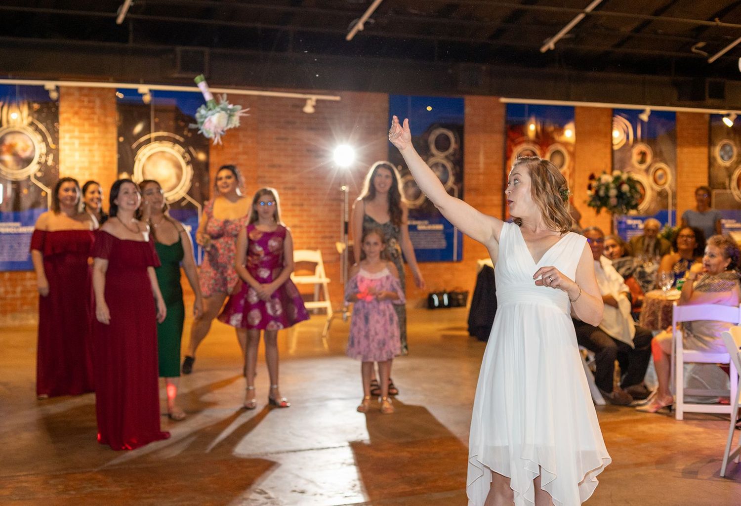 Bride tosses bouquet to excited guests in colorful dresses at a wedding reception in a brick venue.