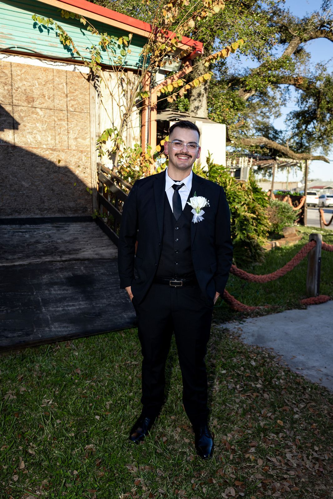 Smiling groom in a black suit with white boutonniere poses outdoors near a rustic building.