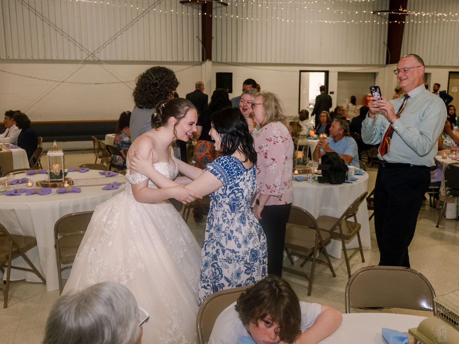 Reception guests interact and dance in a banquet hall decorated with string lights and white linens.