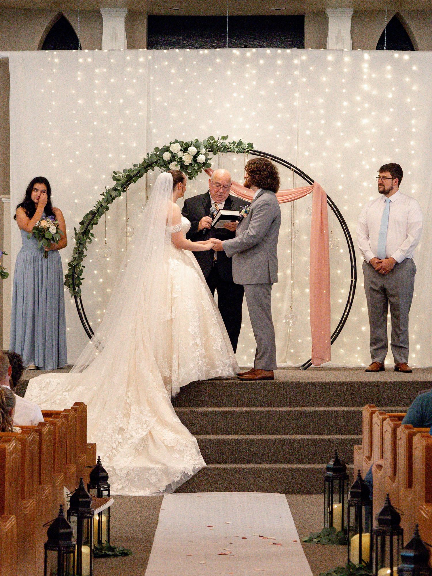 Wedding ceremony unfolds at decorated church altar with string lights and floral arrangements.
