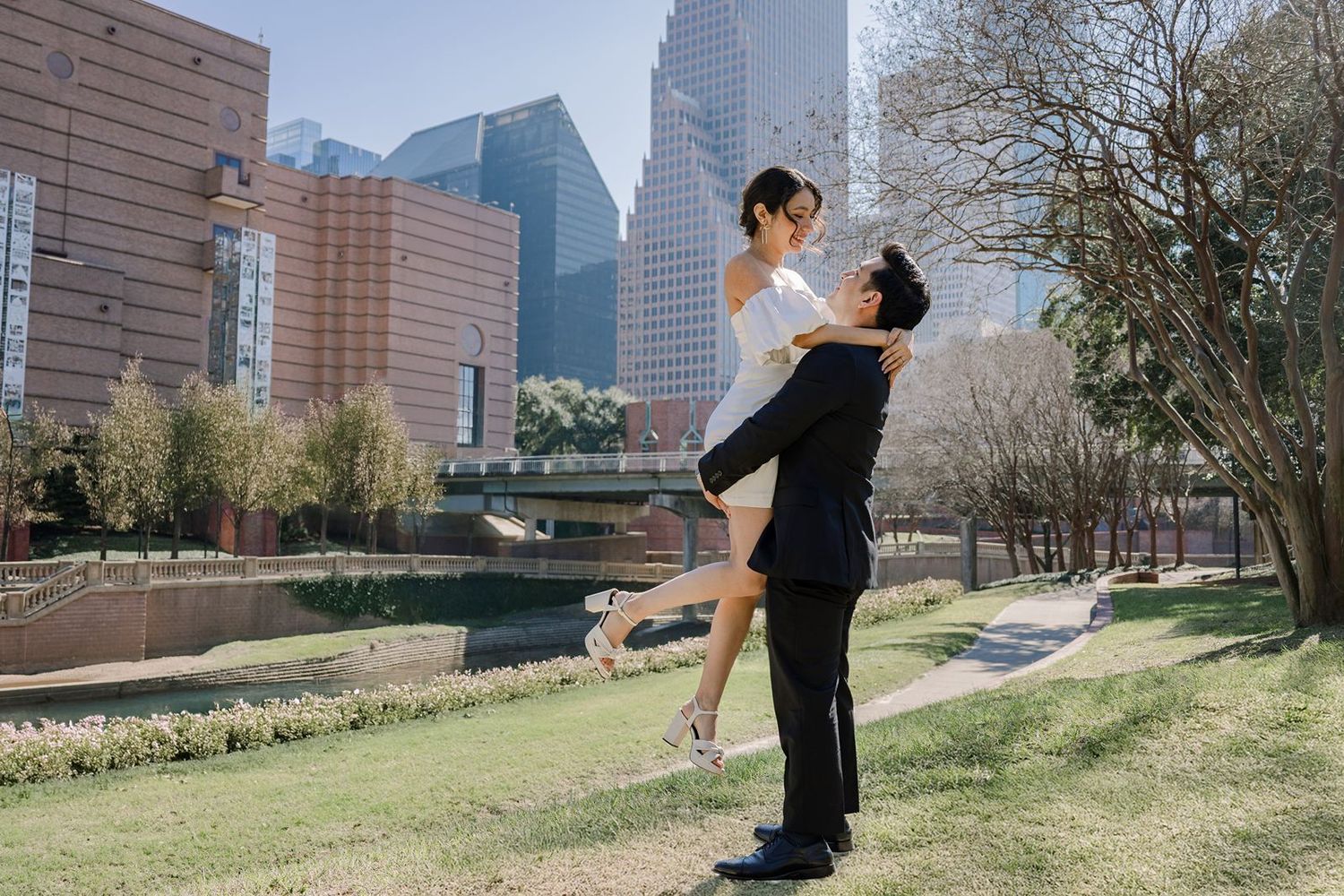 A couple in formal attire dances together in a park with city buildings visible in the background.