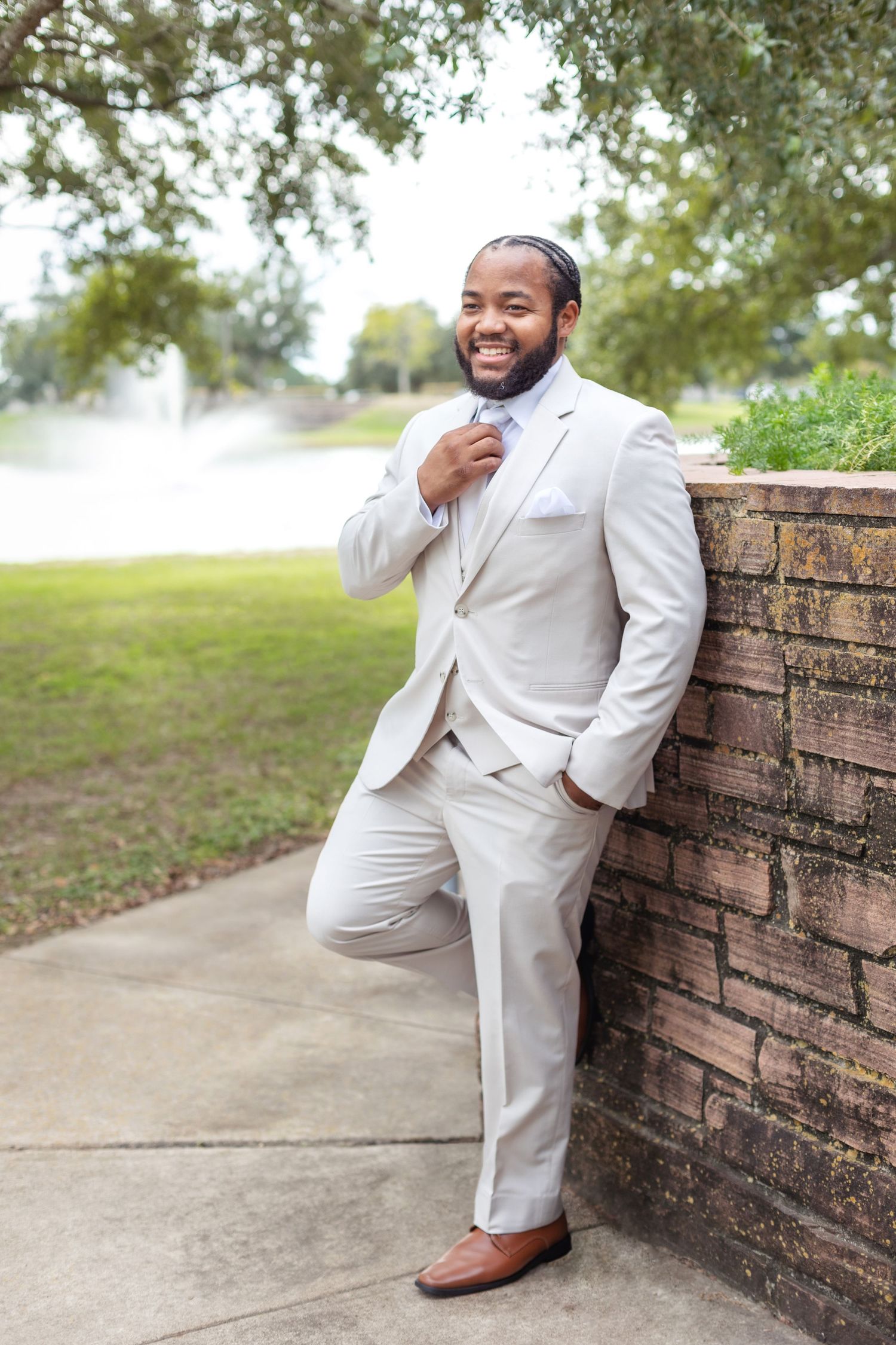 A groom in a light cream colored suit poses playfully against a brick wall outdoors.