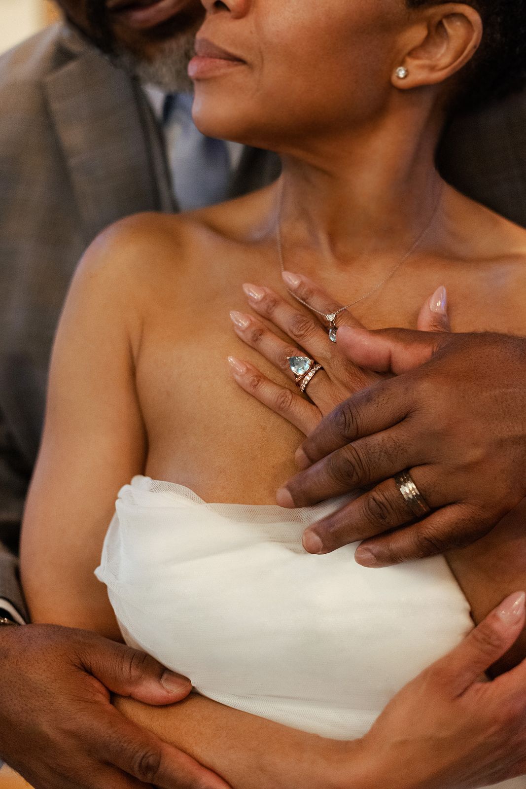 Black couple in wedding attire displaying rings, bride in strapless white gown with blue gemstone engagement ring.