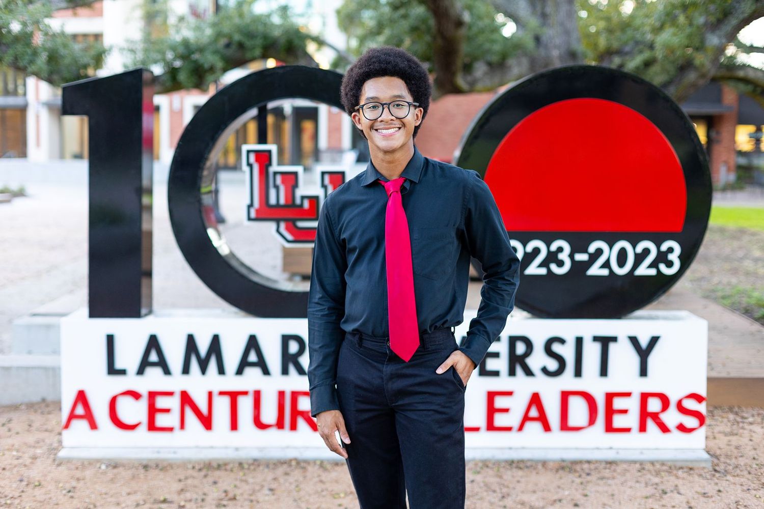 Professional wearing glasses and red tie poses outdoors against blurred university campus background.