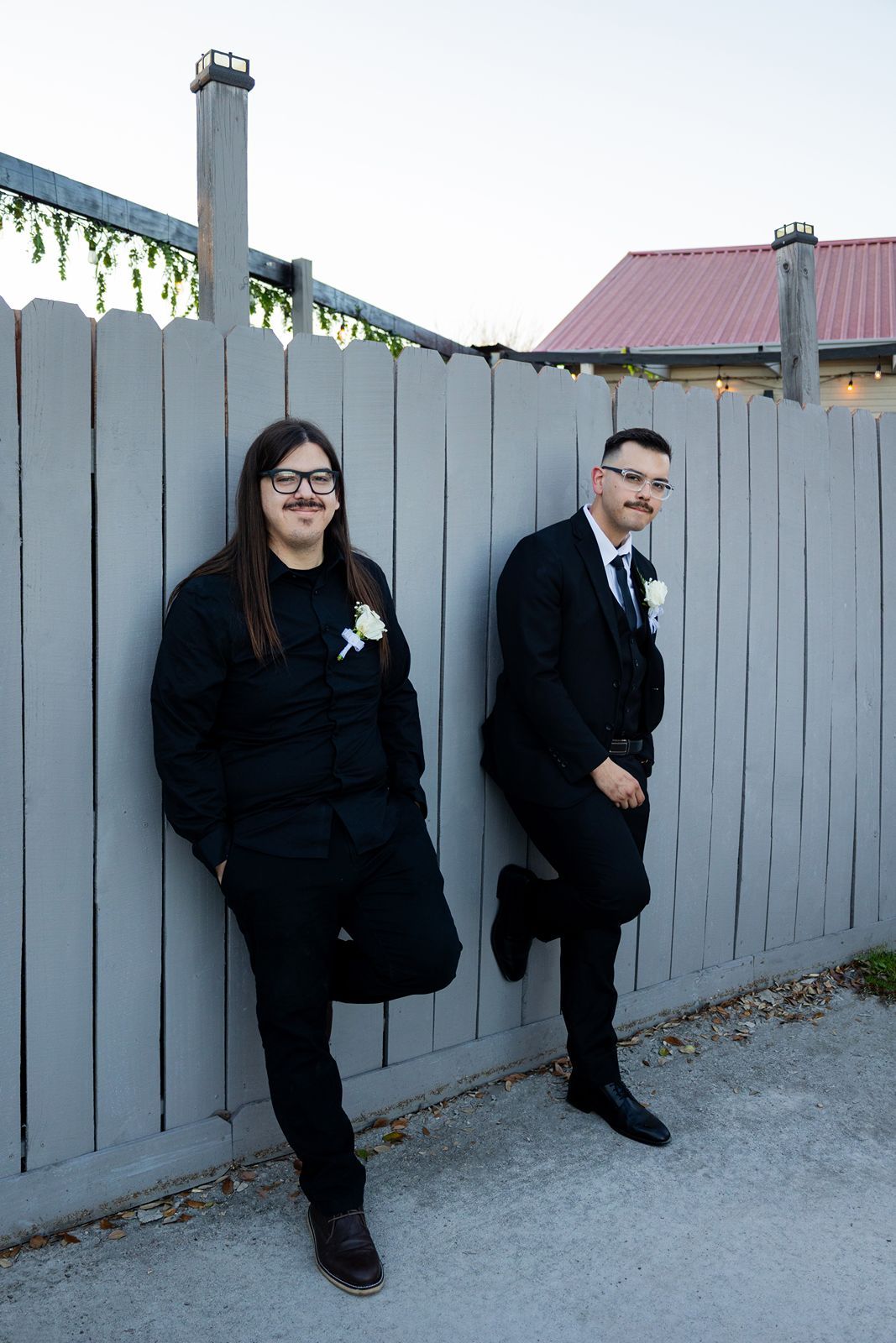Two grooms in black suits with boutonnieres leaning against a gray wooden fence at an outdoor wedding venue.