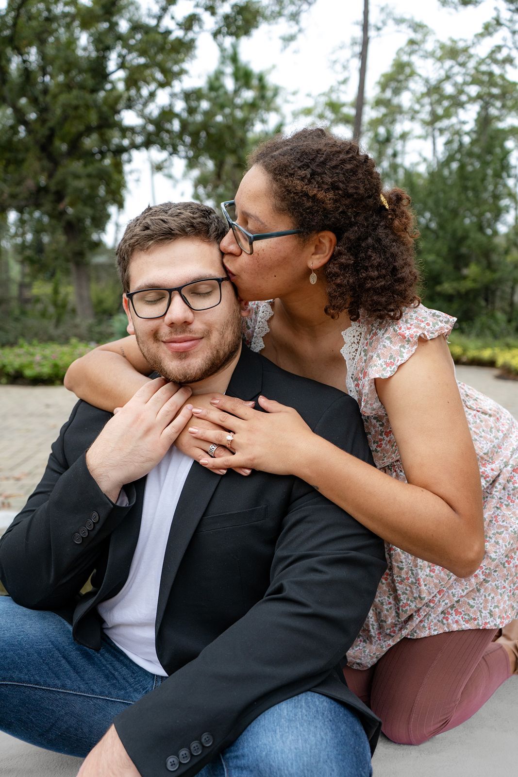 A couple shares a tender moment outdoors, woman kissing mans cheek while embracing him from behind in a garden setting.