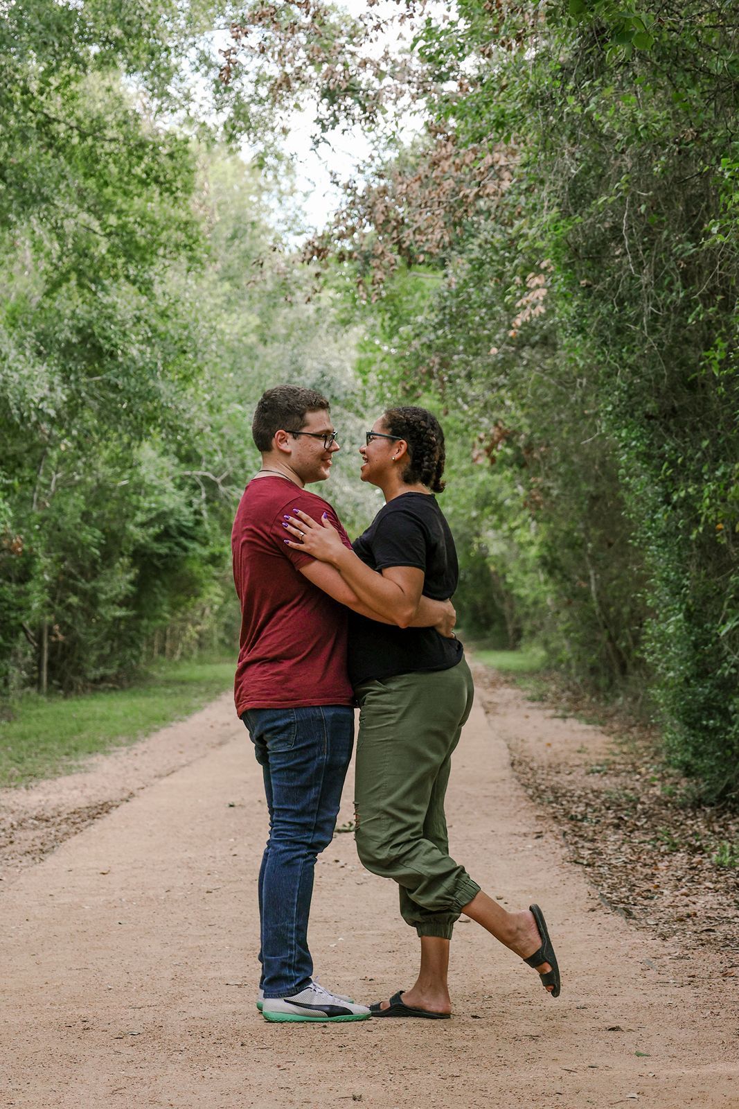 Couple embracing on a tree-lined dirt path, sharing a romantic moment outdoors in a lush green setting.