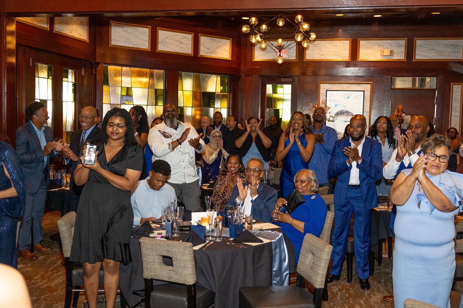 Crowd of elegantly dressed guests applauding and celebrating at a formal indoor banquet event in a wood-paneled venue.