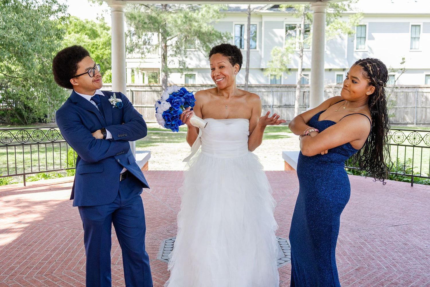 Bride in white gown stands with groomsman and bridesmaid in royal blue, all striking playful poses outdoors.