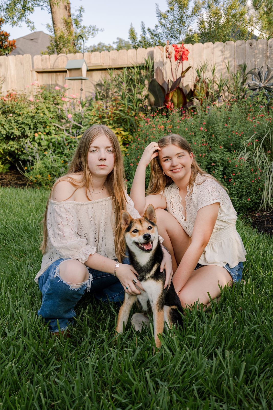 Two people in casual attire sit in tall grass with a happy Shiba Inu mix dog in a garden setting.
