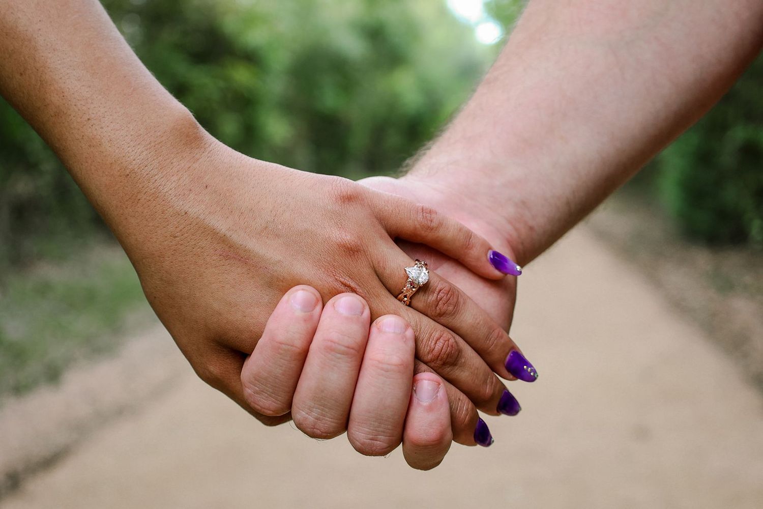 Two people holding hands outdoors, one wearing a diamond engagement ring and the other with purple nail polish.