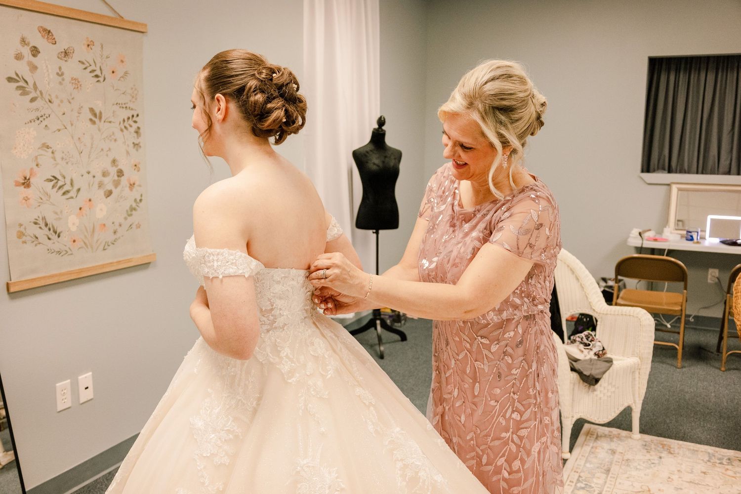 Woman in sparkly rose gold bridesmaid dress assists bride with wedding gown fitting.