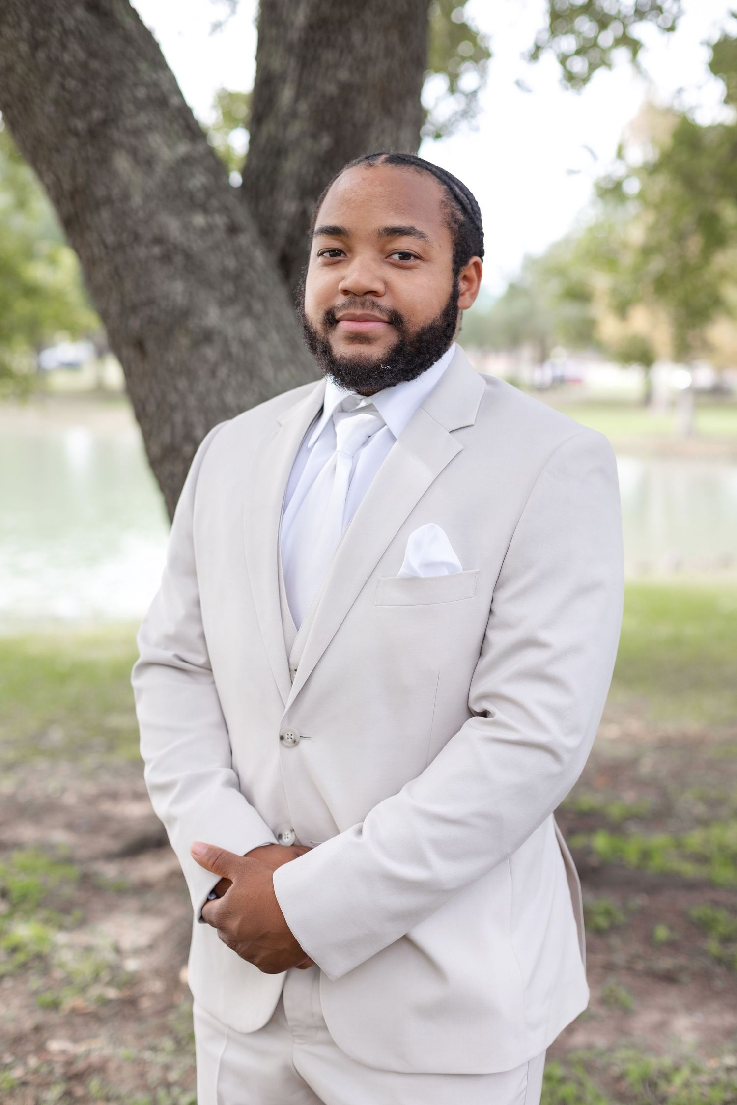 A groom in a crisp white suit and tie poses confidently against a natural outdoor backdrop.