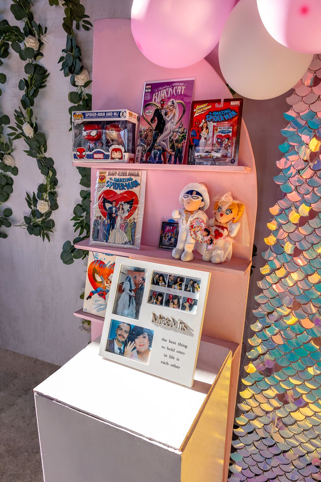 Colorful party display with comic books, plush toys, photo strips, balloons, and iridescent sequin backdrop.