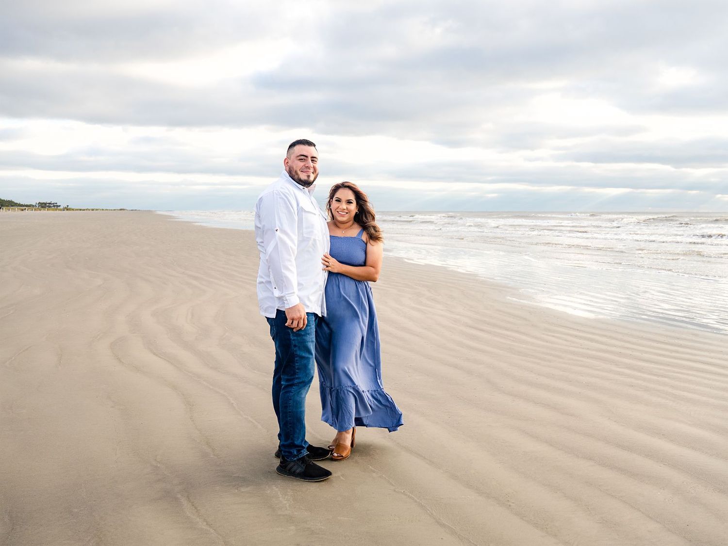 A couple in blue and white outfits walking along a scenic beach with rolling sand dunes and cloudy skies.