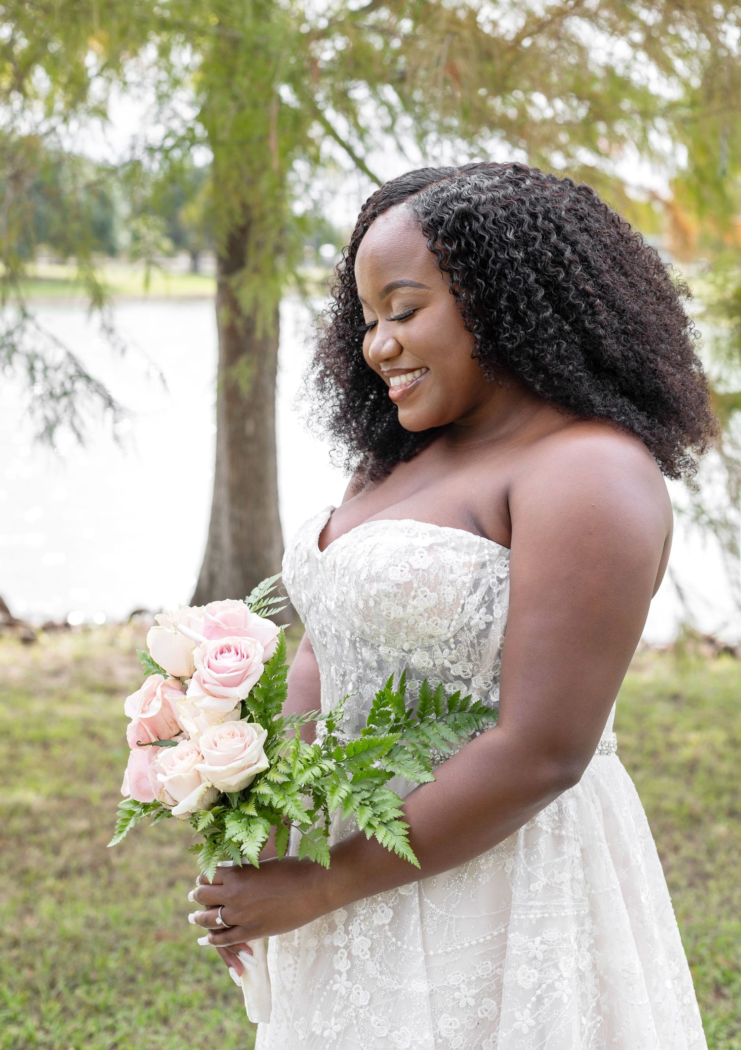 A bride in a lace strapless gown holds delicate pink roses during her outdoor bridal portraits.