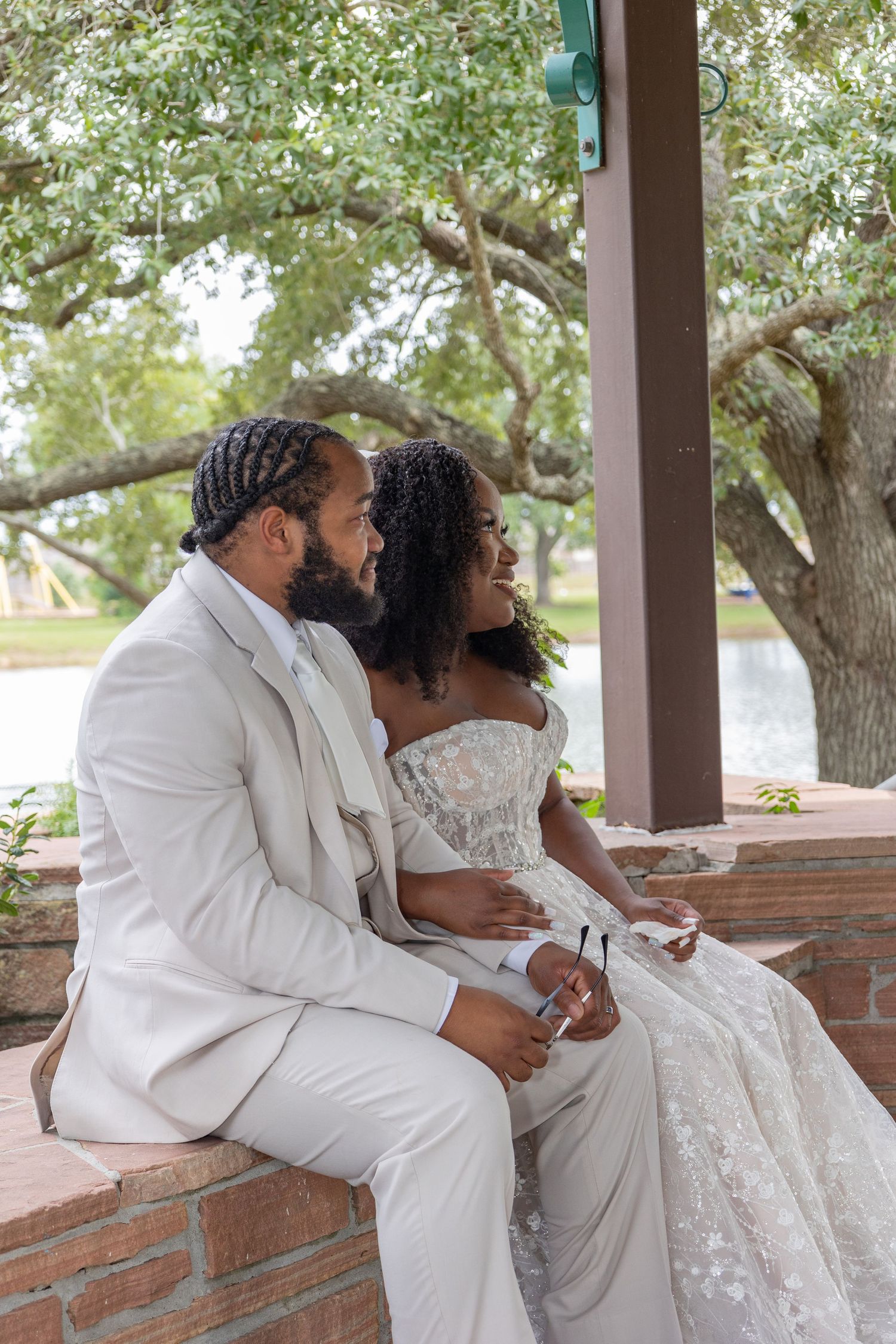 A couple in white wedding attire sits close together on a brick wall during an outdoor celebration.