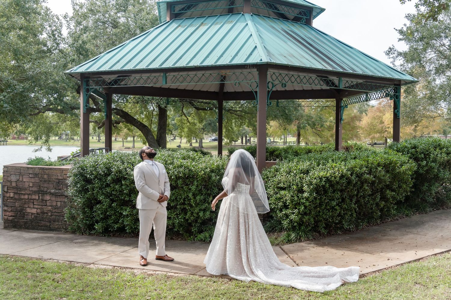 A couple shares a first look moment under a green-roofed gazebo surrounded by lush landscaping.