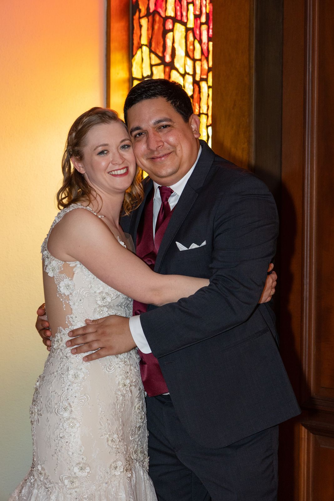 Smiling bride and groom embrace warmly beside a stained glass window at their wedding reception.