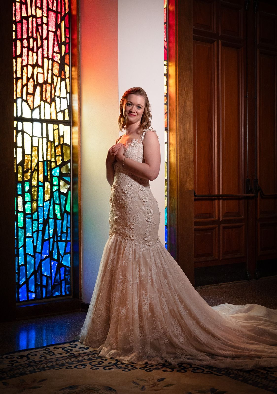 A bride poses in a lace mermaid wedding gown next to a colorful stained glass window in a church.
