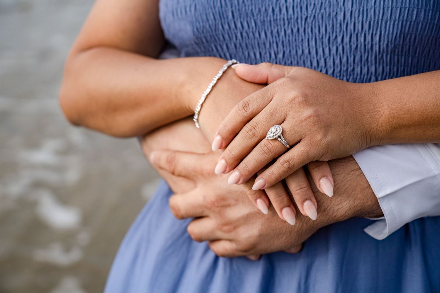 Close-up of hands wearing engagement ring and bracelet with light blue dress against blurred background.