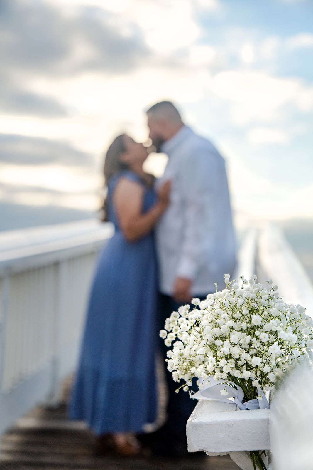 White baby's breath flowers in soft focus with a couple embracing on a pier in the background.
