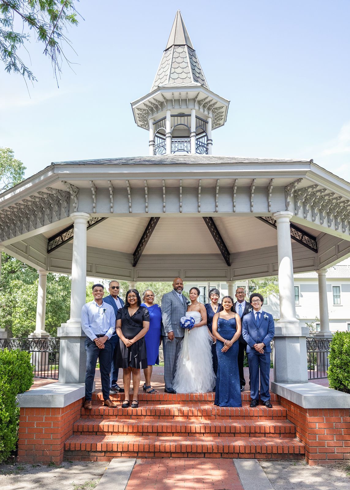 Wedding party group photo on brick steps of white Victorian gazebo with family dressed in blue.