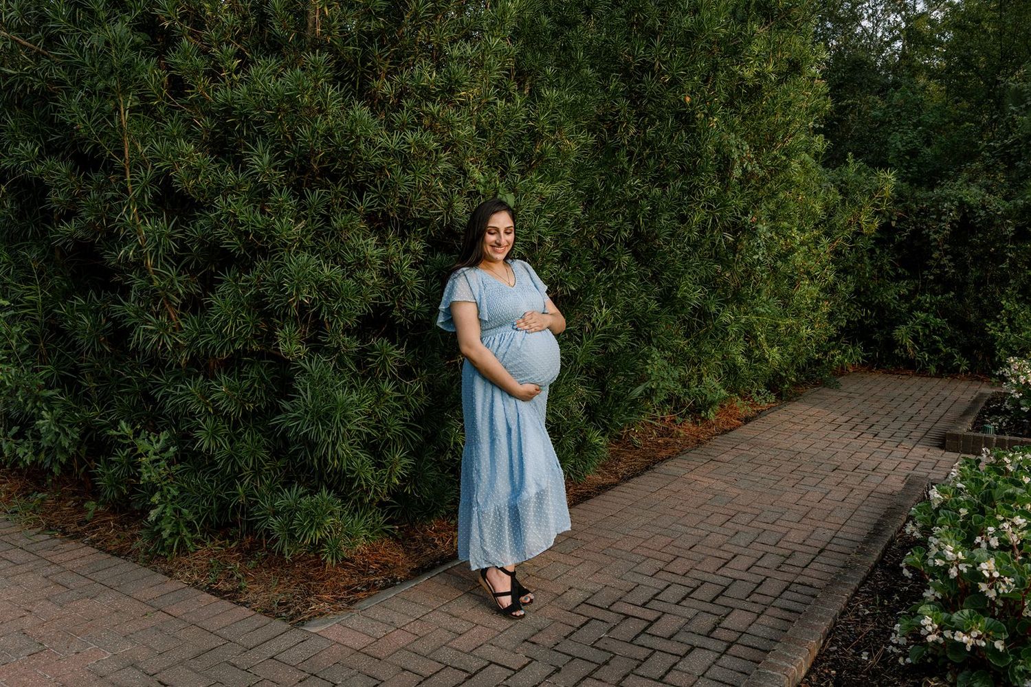 A person in a light blue dress poses in different positions along a brick pathway surrounded by lush greenery.