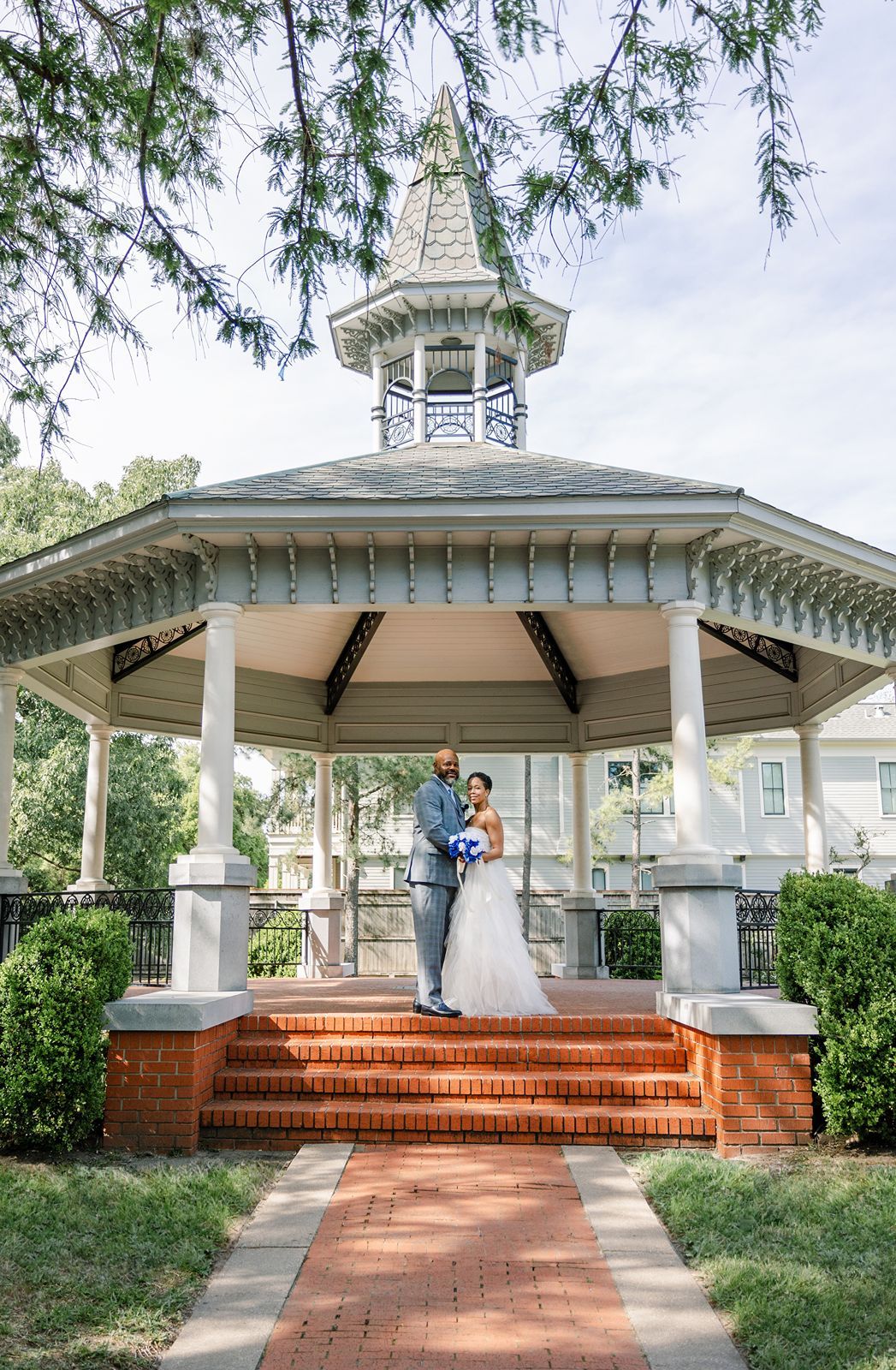 A couple in wedding attire poses on brick steps of a charming gazebo surrounded by lush greenery.