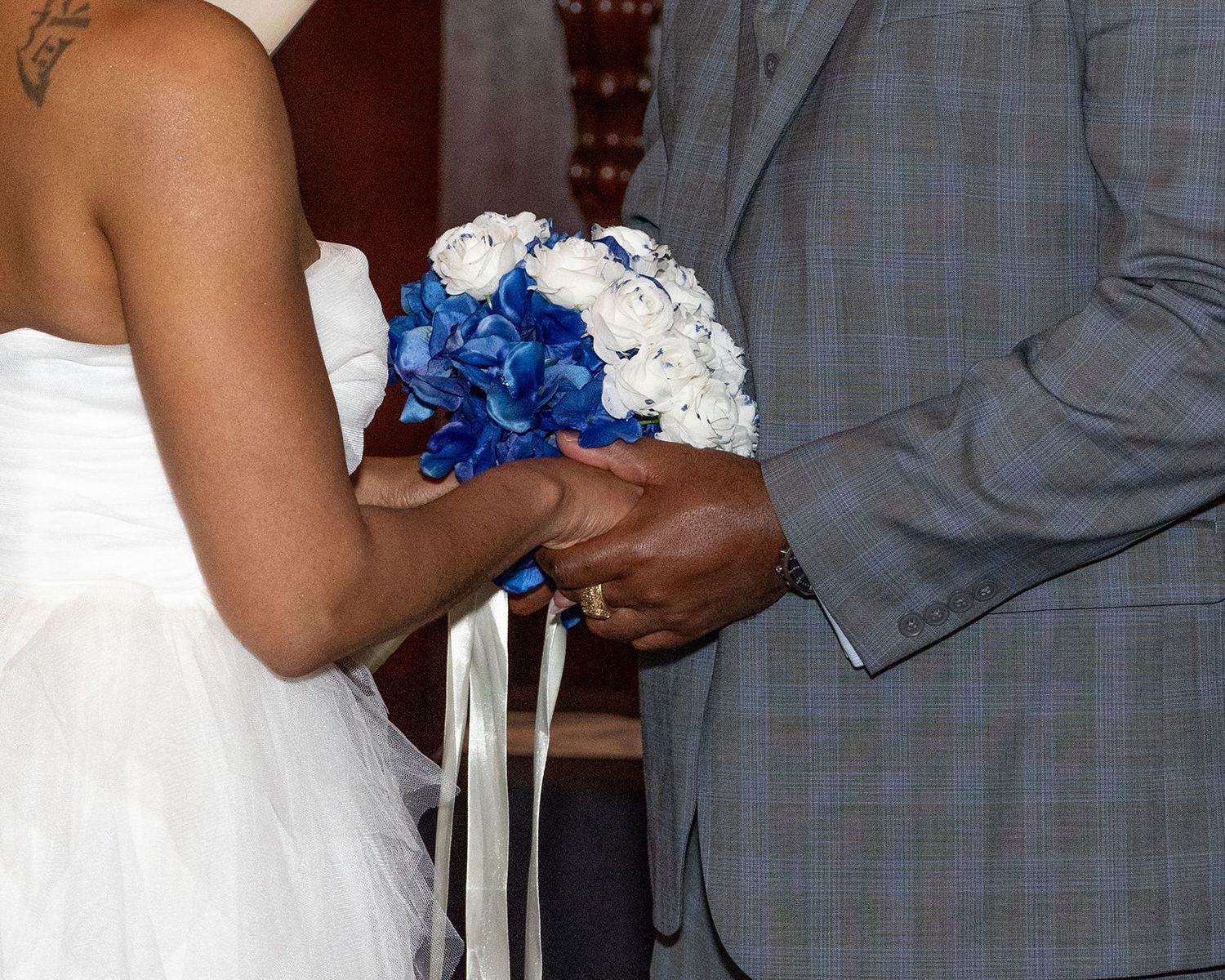 Bride and groom holding blue and white wedding bouquet during ceremony, bride in white dress, groom in plaid suit.