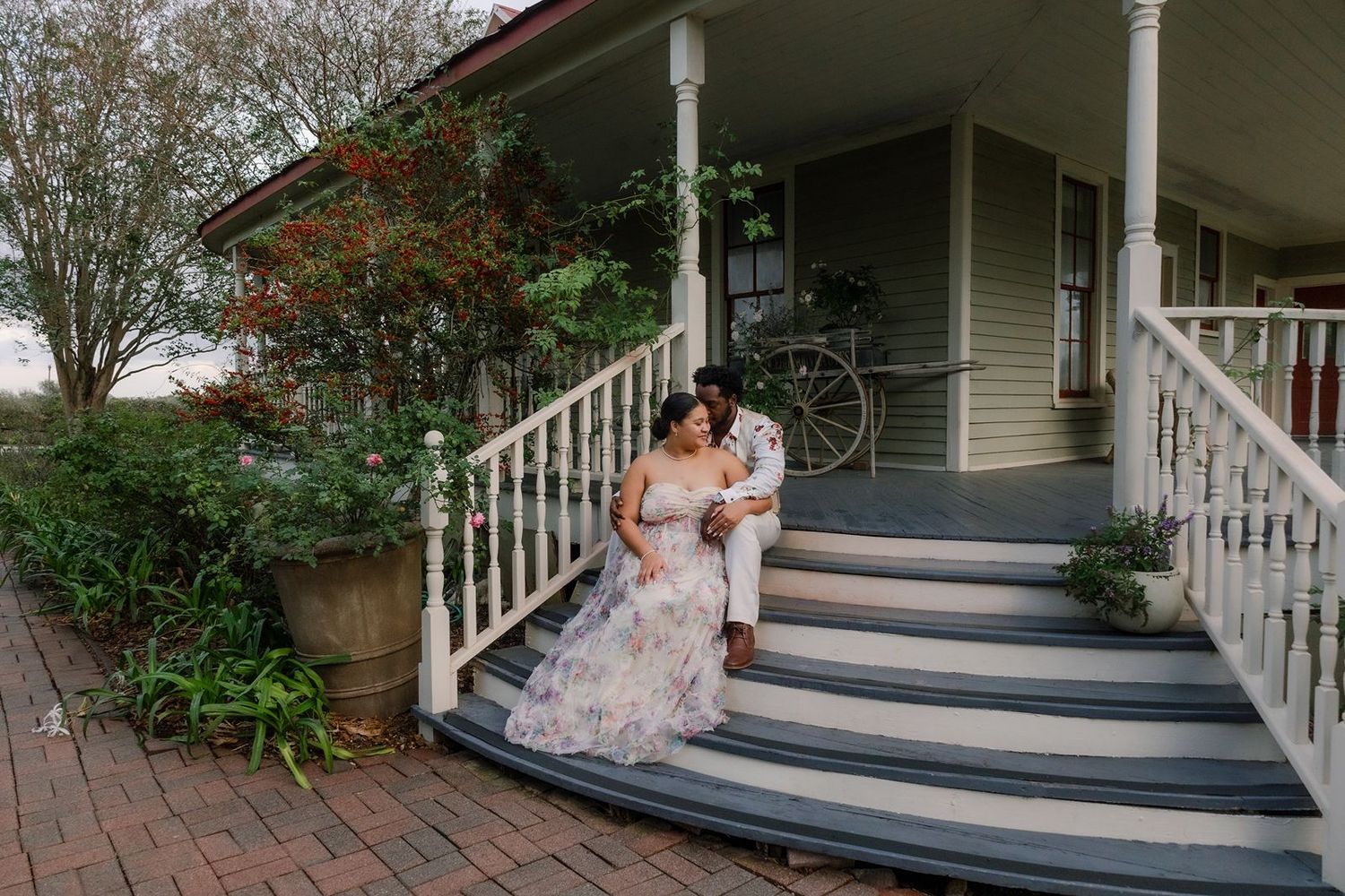 A couple in wedding attire sits together on the steps of a charming white-railed porch of a vintage house.