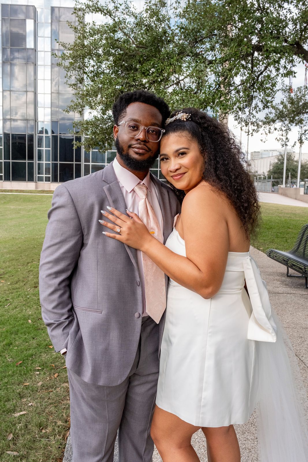 A couple in formal attire sharing romantic moments against an urban backdrop with glass buildings and trees.