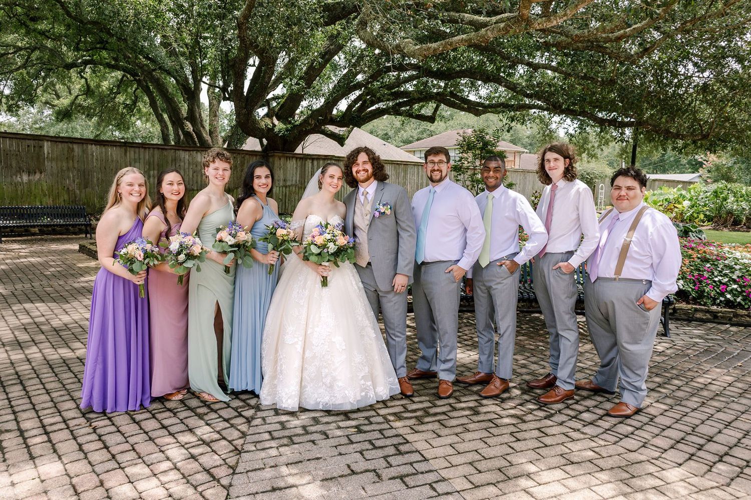 Wedding party in coordinated purple and gray attire poses together on a brick pathway in a garden setting.