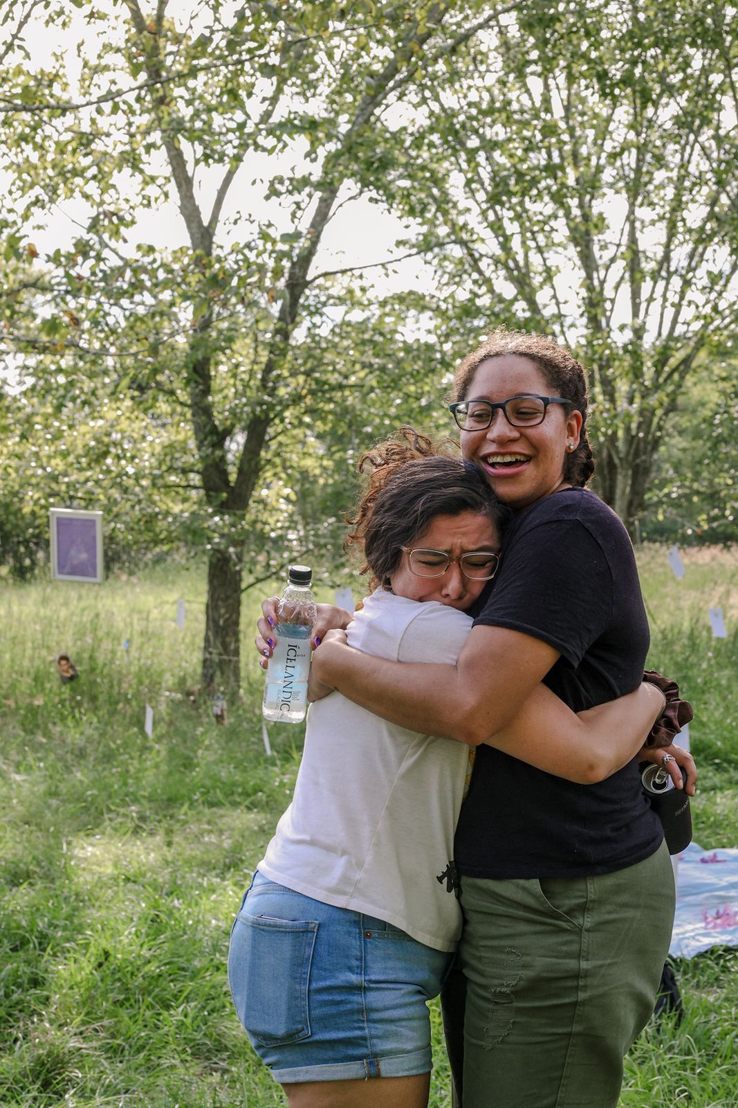 Two women hugging and celebrating outdoors in a grassy field, one crying with joy holding a bottle.