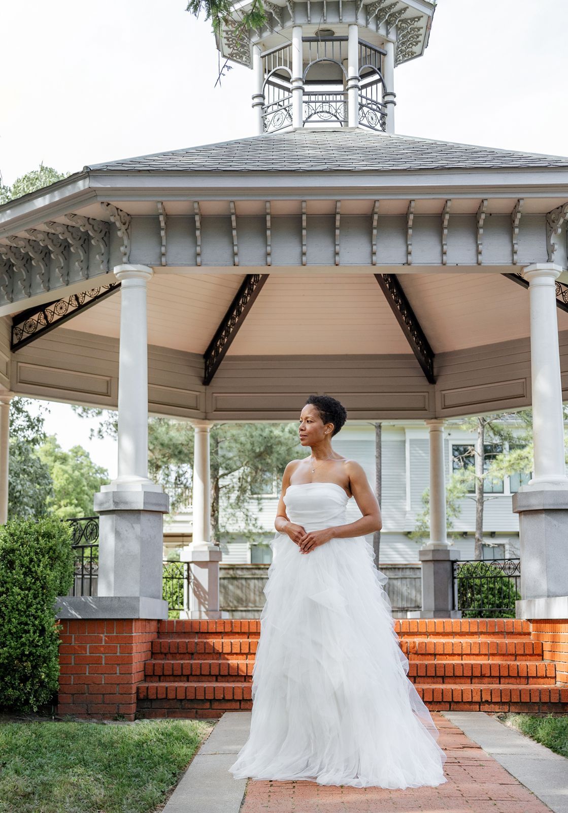 A bride in a white wedding dress poses gracefully in front of a white gazebo with red brick steps.