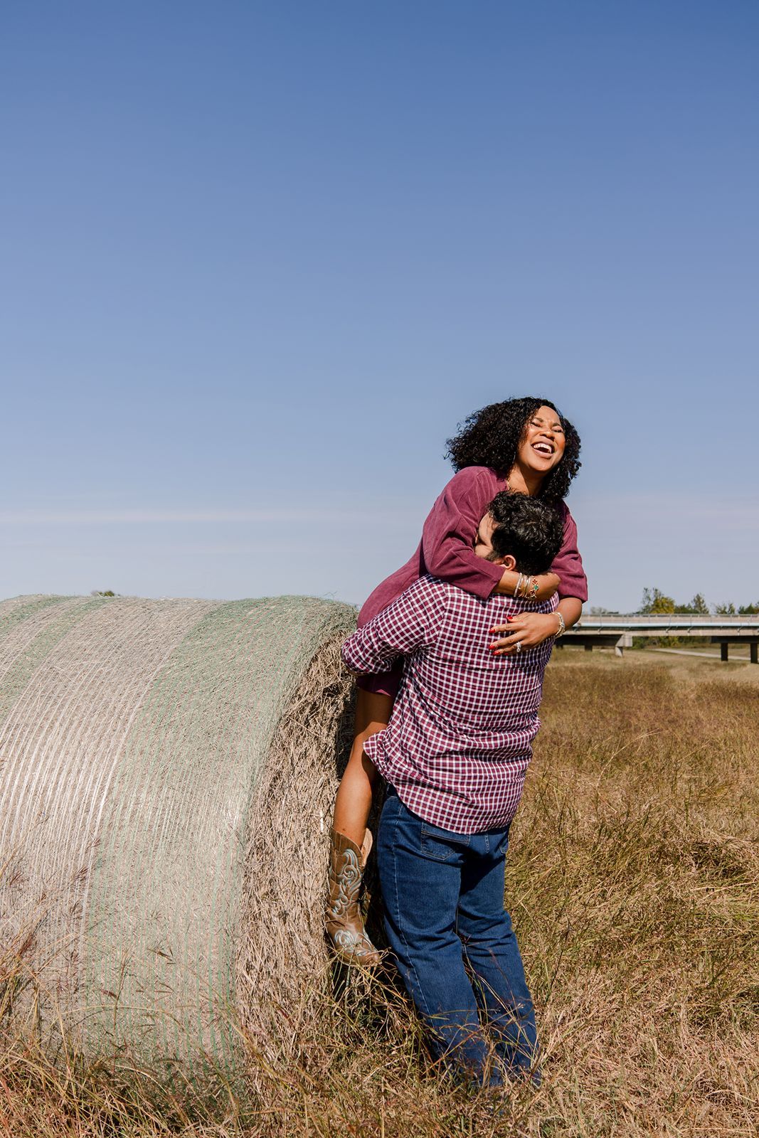 Couple sharing playful moments together while sitting against round hay bales in a sunny field.