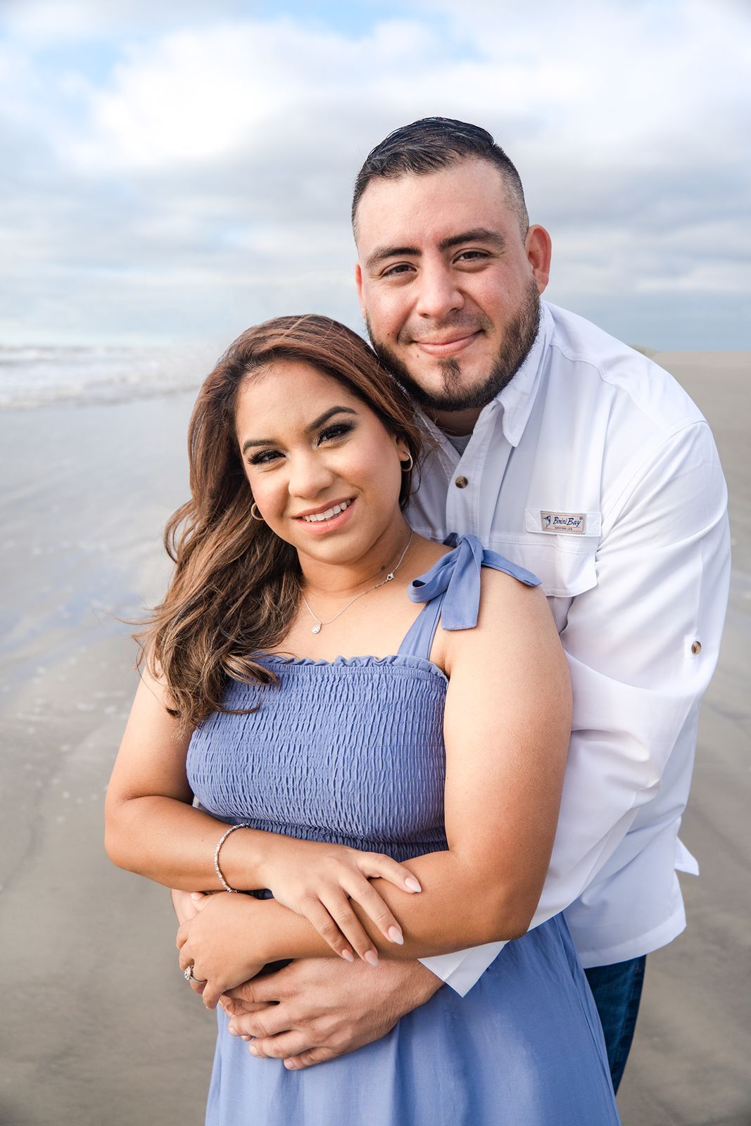 A couple shares romantic moments together at the beach on a cloudy day, wearing blue and white outfits.