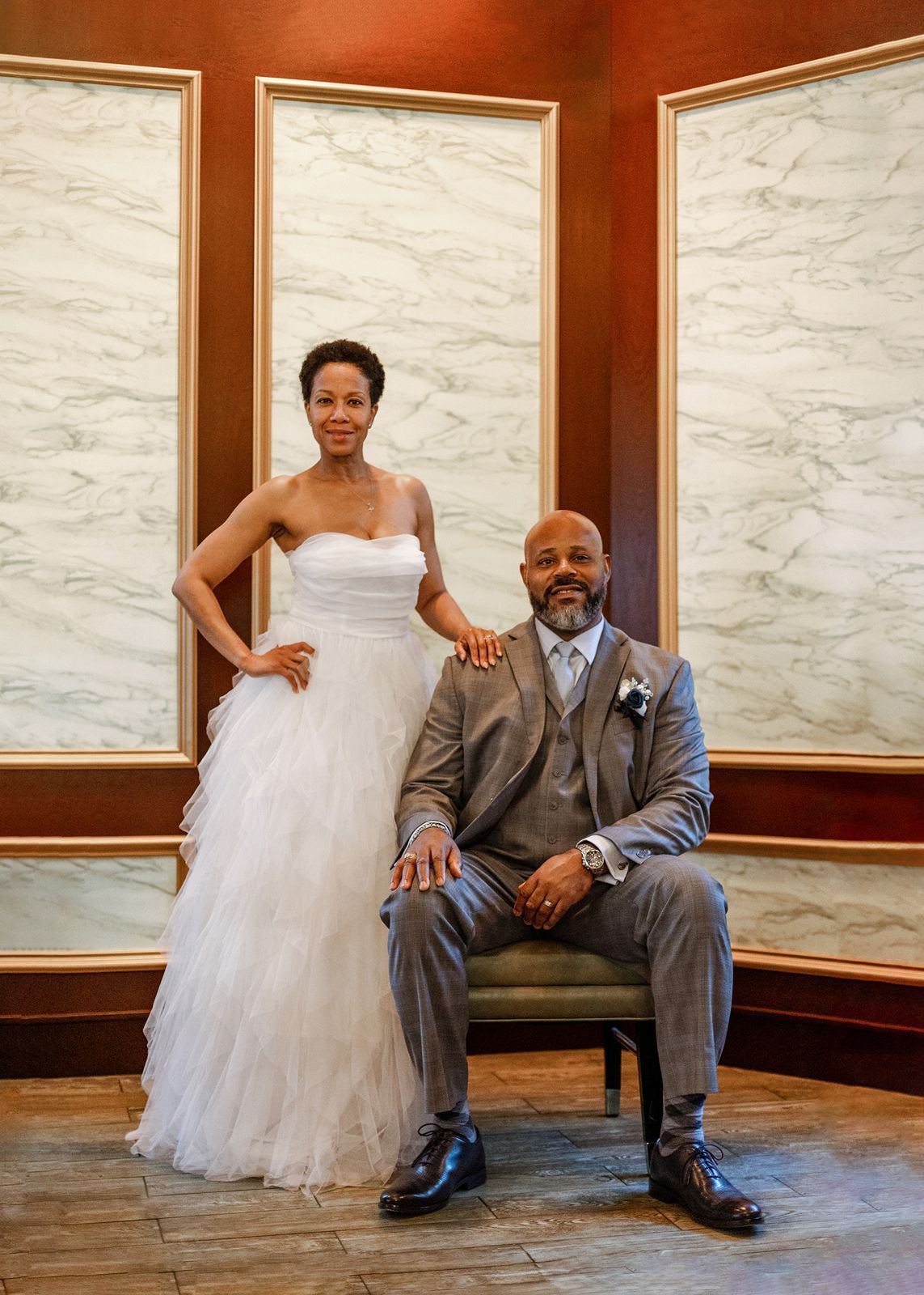 Elegant wedding couple posing in marble-walled venue, bride in white strapless gown, groom seated in gray suit.