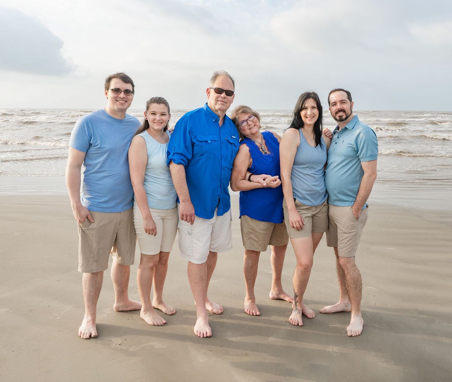 A group of people wearing coordinated blue and gray outfits pose together on a sandy beach at sunset.
