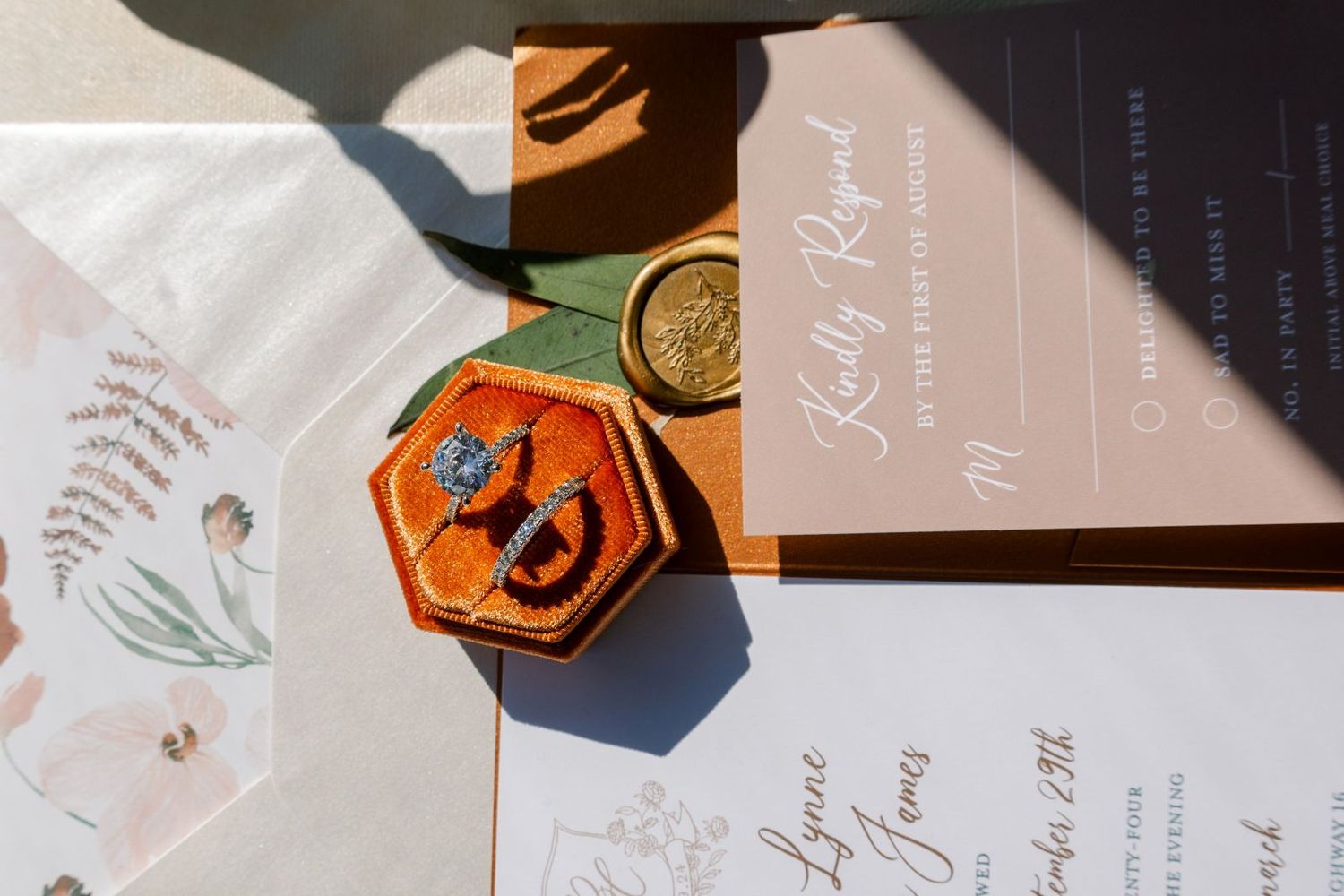 Wedding stationery flatlay with his and hers wedding bands, calligraphy invitations, and envelopes in warm fall tones.