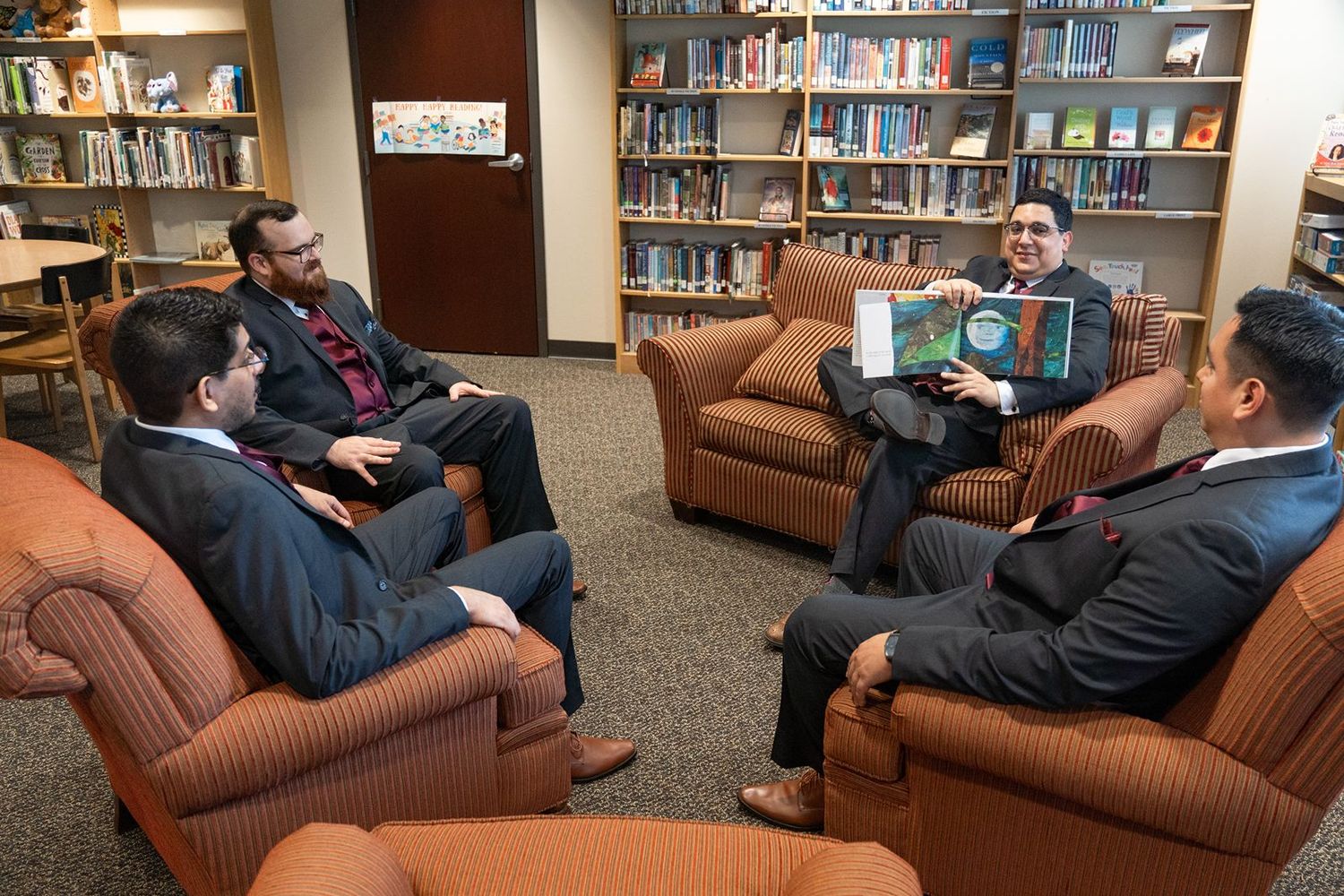 Four men in suits discuss a book in a library seating area with bookshelves behind them.