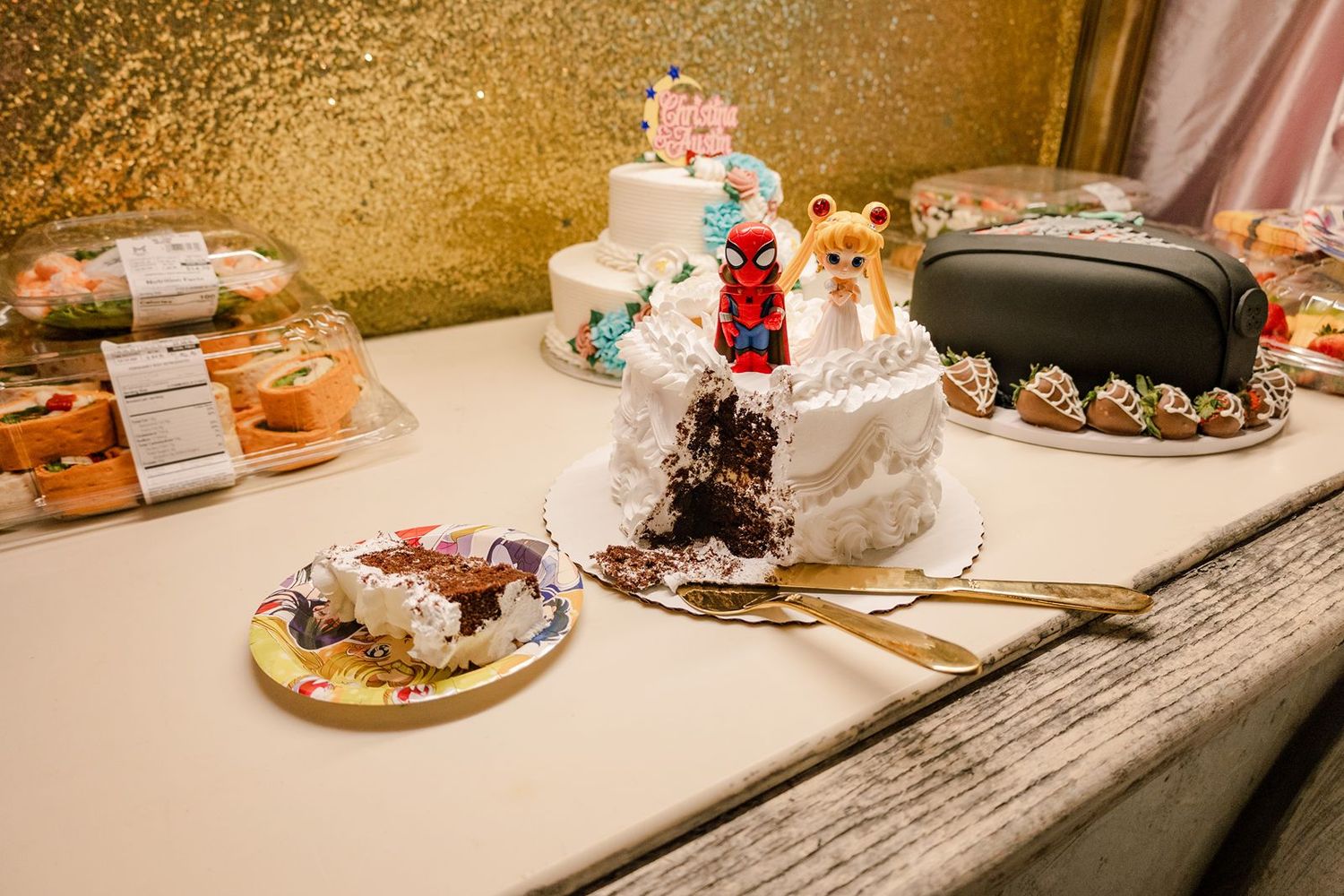 Decorated wedding cakes on a white table, including a cut cake revealing chocolate interior, with gold serving utensils.