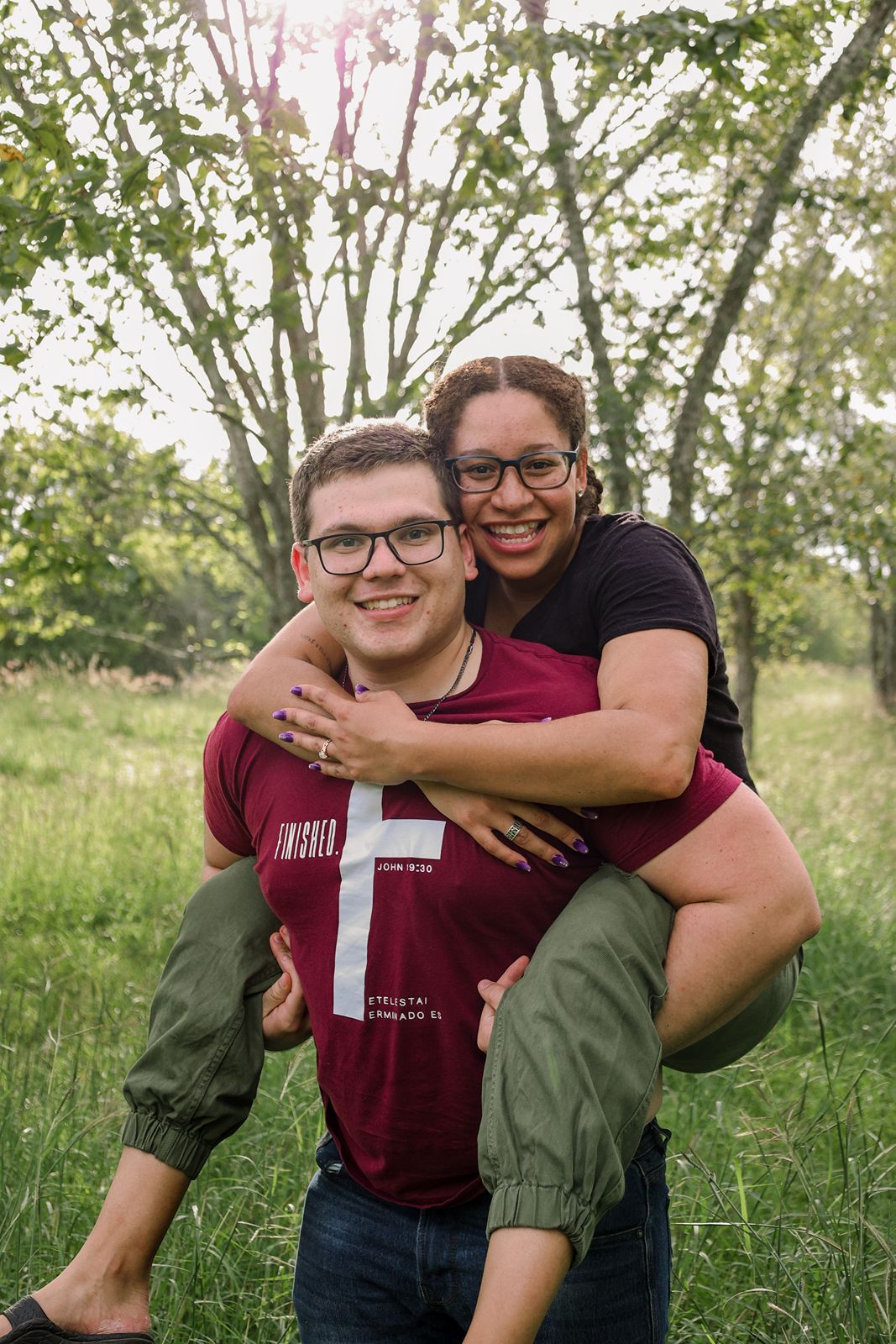 A smiling couple poses outdoors, man giving woman a piggyback ride in a green park setting.