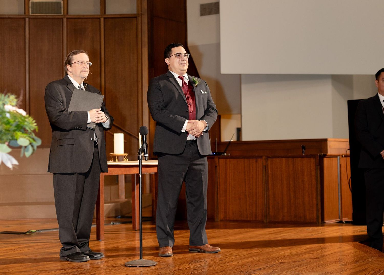 Two men in suits stand at a church altar podium during a wedding ceremony.