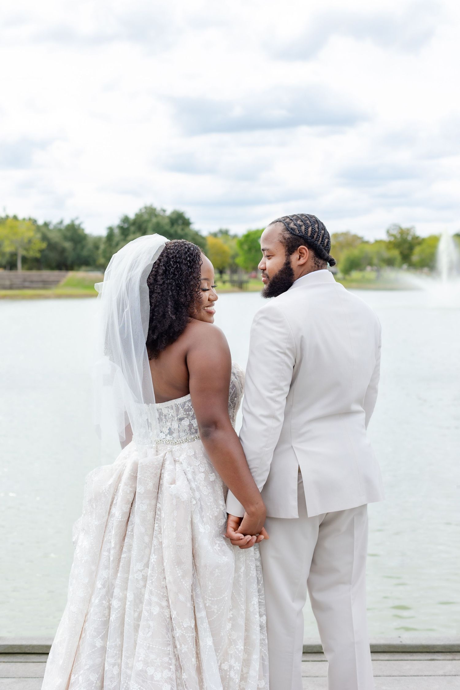 A wedding couple holds hands while walking along a lakeside dock in formal white attire.