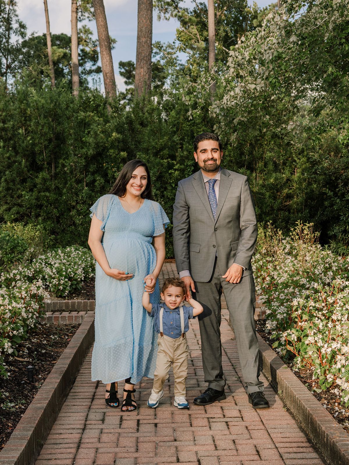 A family stands together on a brick pathway surrounded by lush greenery and flowering bushes in a garden setting.